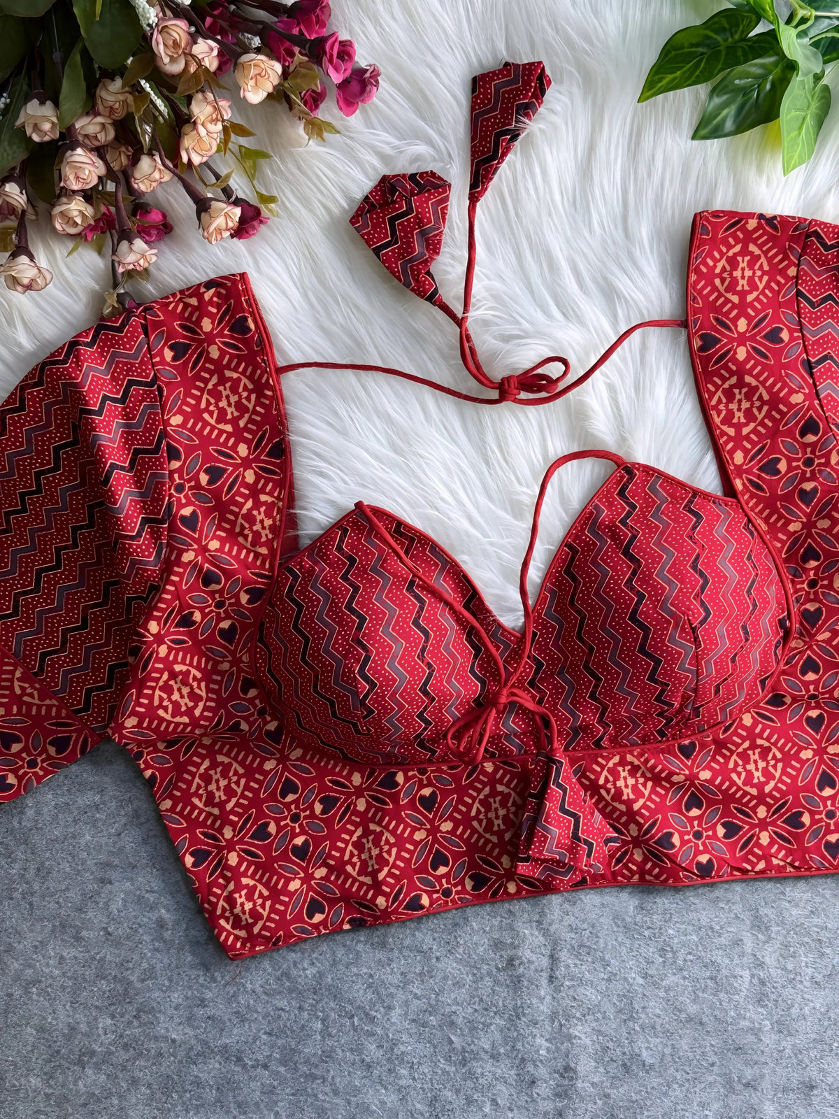 Red printed padded blouse with geometric and floral patterns, displayed on a white fur surface with artificial flowers and green leaves in the background.