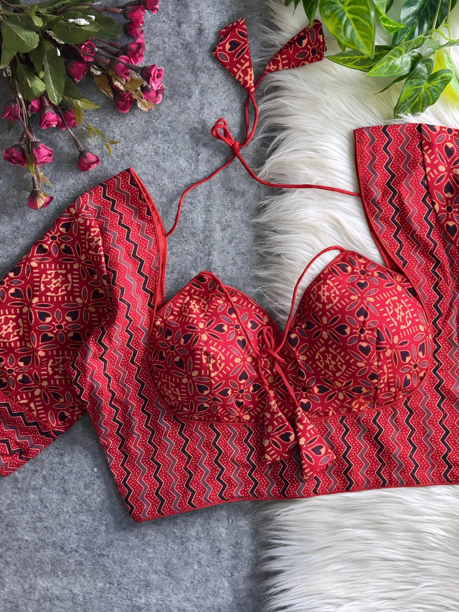 Red printed padded blouse with geometric and floral patterns, displayed on a white fur surface with artificial flowers and green leaves in the background.