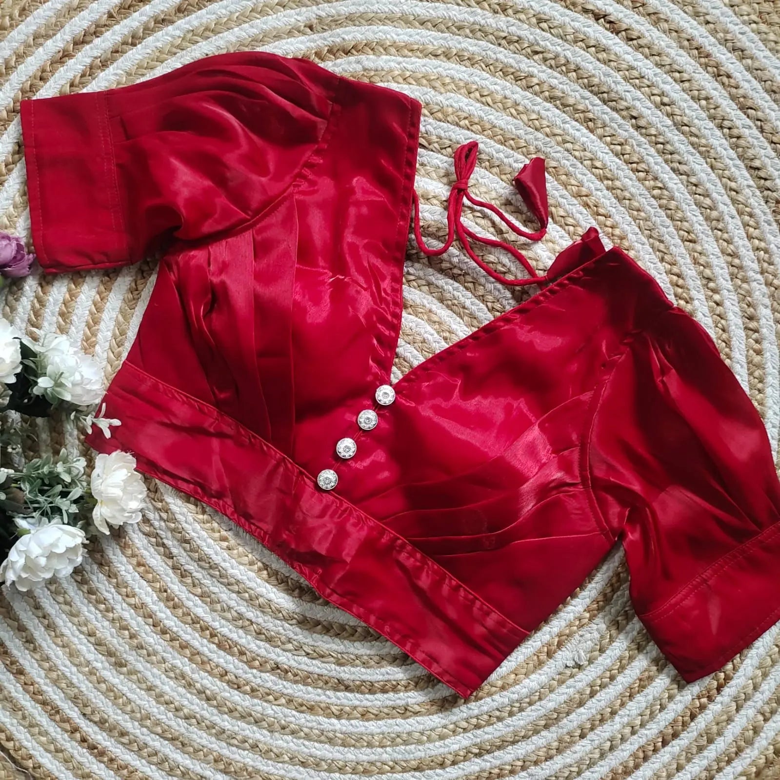 Red satin blouse with pleated detailing and four decorative silver buttons on the front, featuring elbow-length sleeves and tie-up tassels, displayed on a circular woven mat with white flowers on the side.