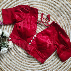 Red satin blouse with pleated detailing and four decorative silver buttons on the front, featuring elbow-length sleeves and tie-up tassels, displayed on a circular woven mat with white flowers on the side.