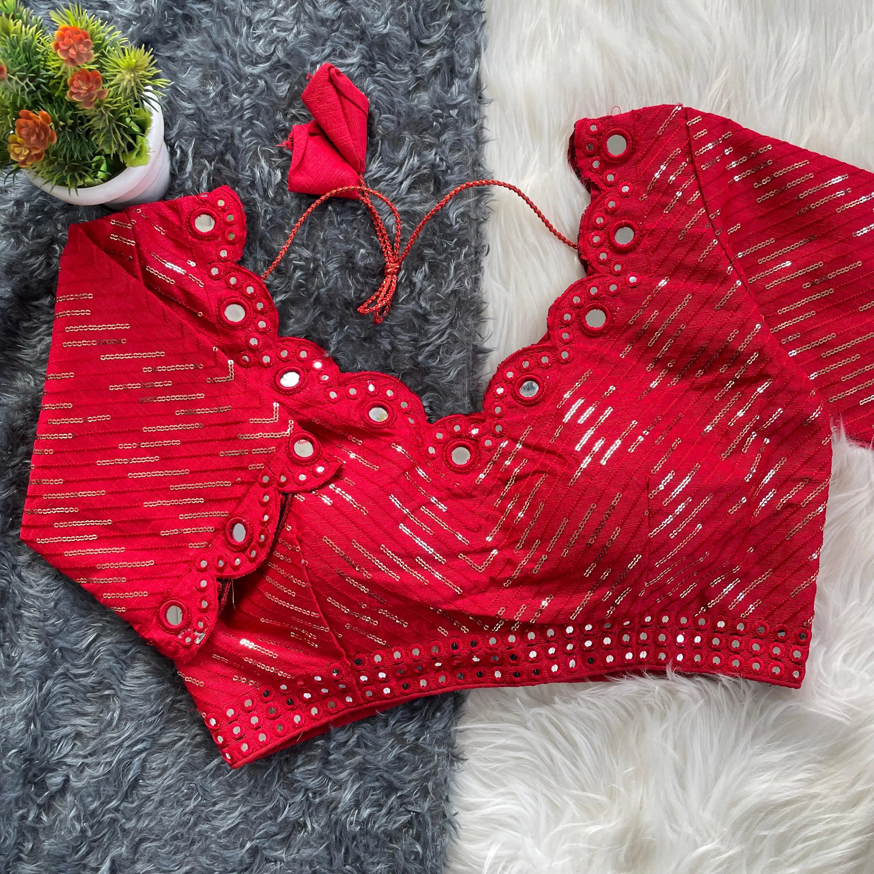 Red bridal blouse with scallop sleeves, featuring silver sequin stripes and prominent round mirror work along the neckline, sleeves, and hem, displayed on a textured grey and white fur background with a small potted plant nearby.