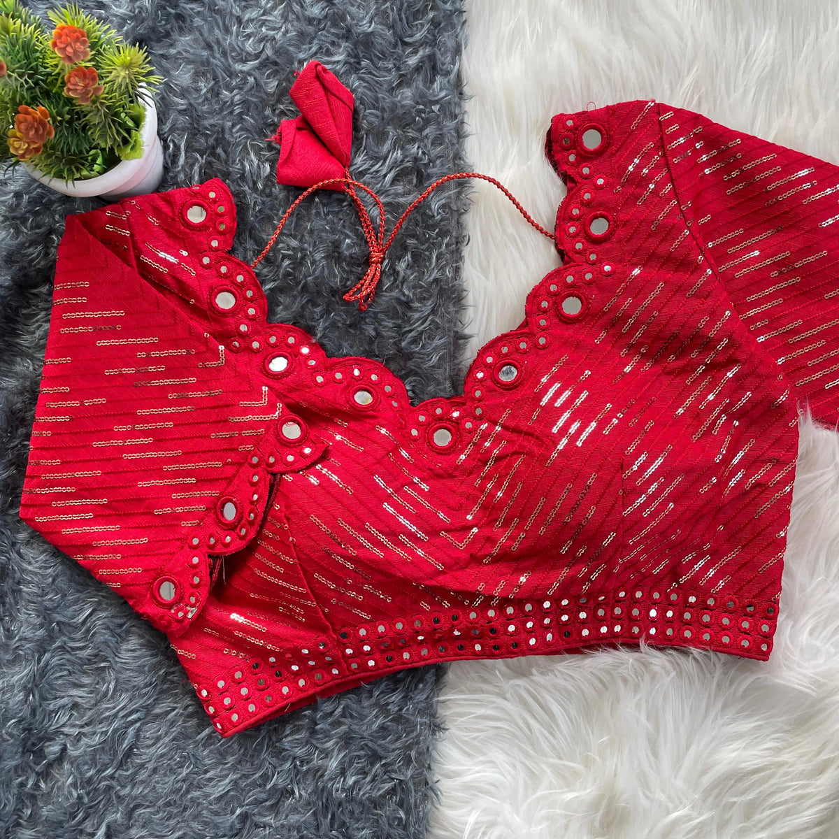 Red bridal blouse with scallop sleeves, featuring silver sequin stripes and prominent round mirror work along the neckline, sleeves, and hem, displayed on a textured grey and white fur background with a small potted plant nearby.