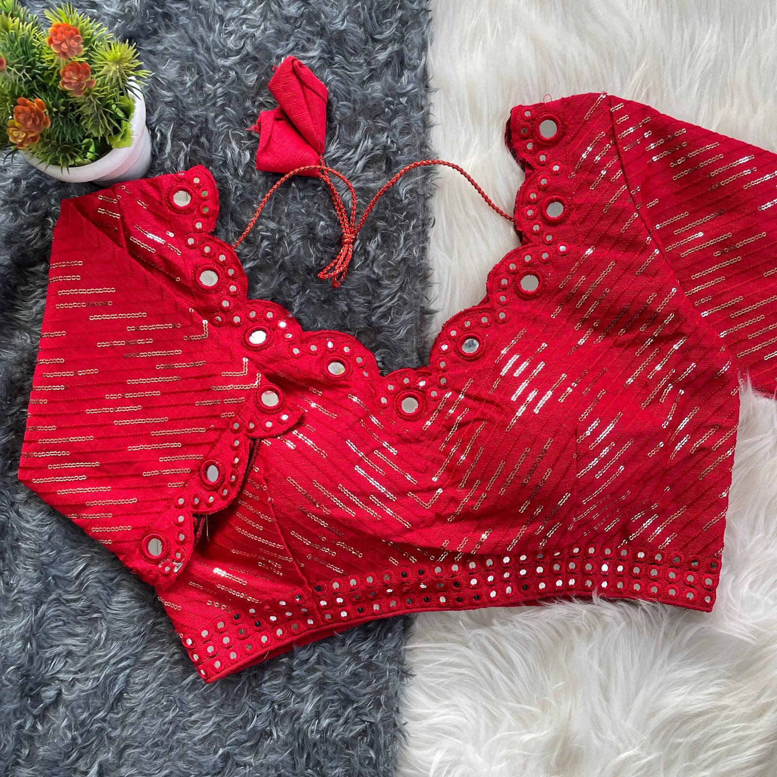 Red bridal blouse with scallop sleeves, featuring silver sequin stripes and prominent round mirror work along the neckline, sleeves, and hem, displayed on a textured grey and white fur background with a small potted plant nearby.