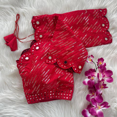 Red bridal blouse with scallop sleeves, featuring silver sequin stripes and prominent round mirror work along the neckline, sleeves, and hem, displayed on a textured grey and white fur background with a small potted plant nearby.