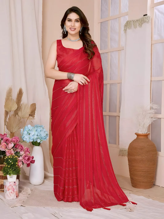 A woman models a vibrant Crimson Red Rimzim Silk Saree. The sheer fabric features prominent vertical stripes and scattered embellishments of fine metallic (silver) threadwork. She wears a matching sleeveless red blouse and accessorizes with chunky oxidized silver jewelry, including a layered necklace and a wide cuff bangle, posing indoors.