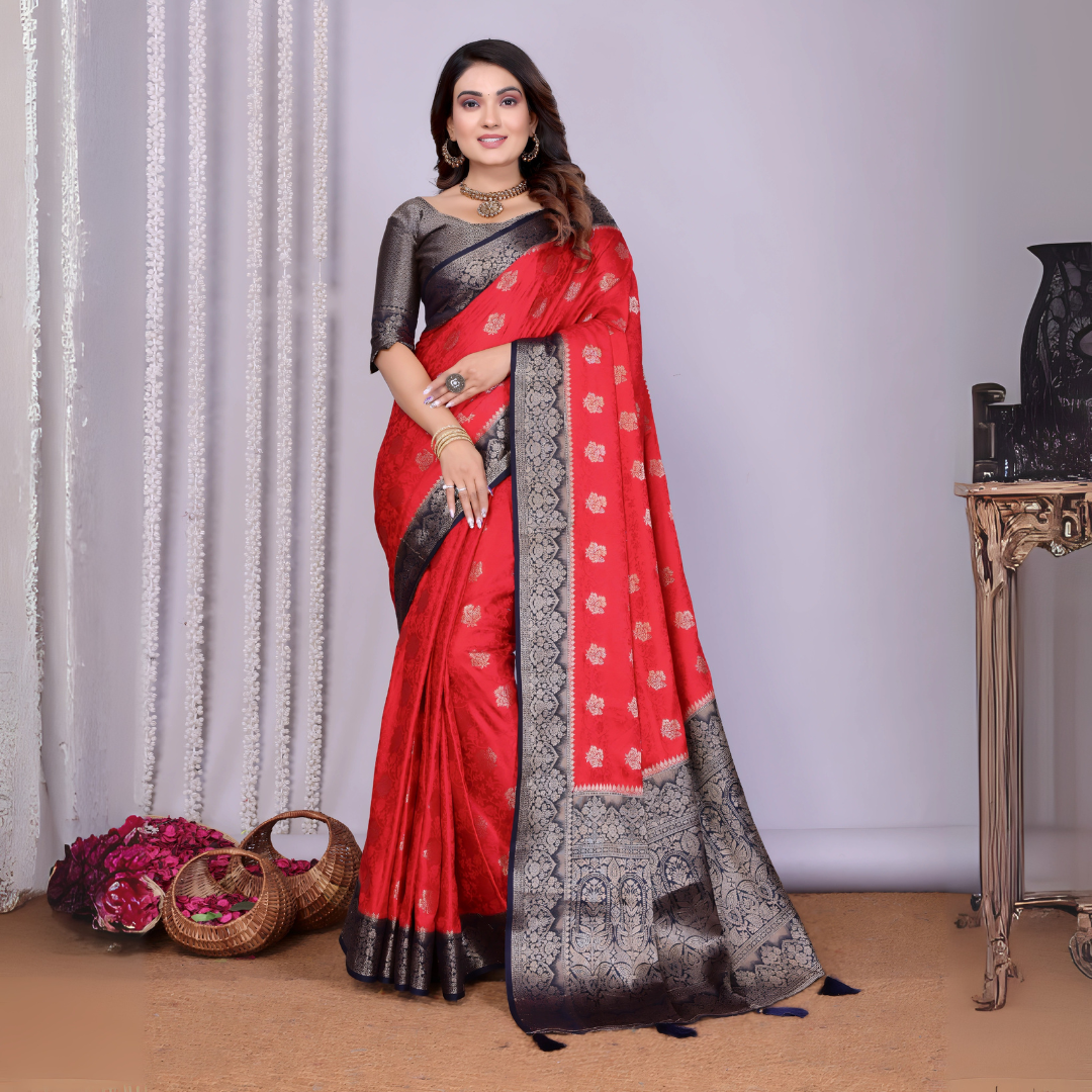 Woman wearing a striking red silk saree with intricate silver and black zari border, detailed buti motifs, and a matching black blouse. She is accessorized with traditional jewelry and bangles, posing elegantly in a contemporary studio setting with woven baskets of rose petals, sheer white curtains, and a neutral gray backdrop.