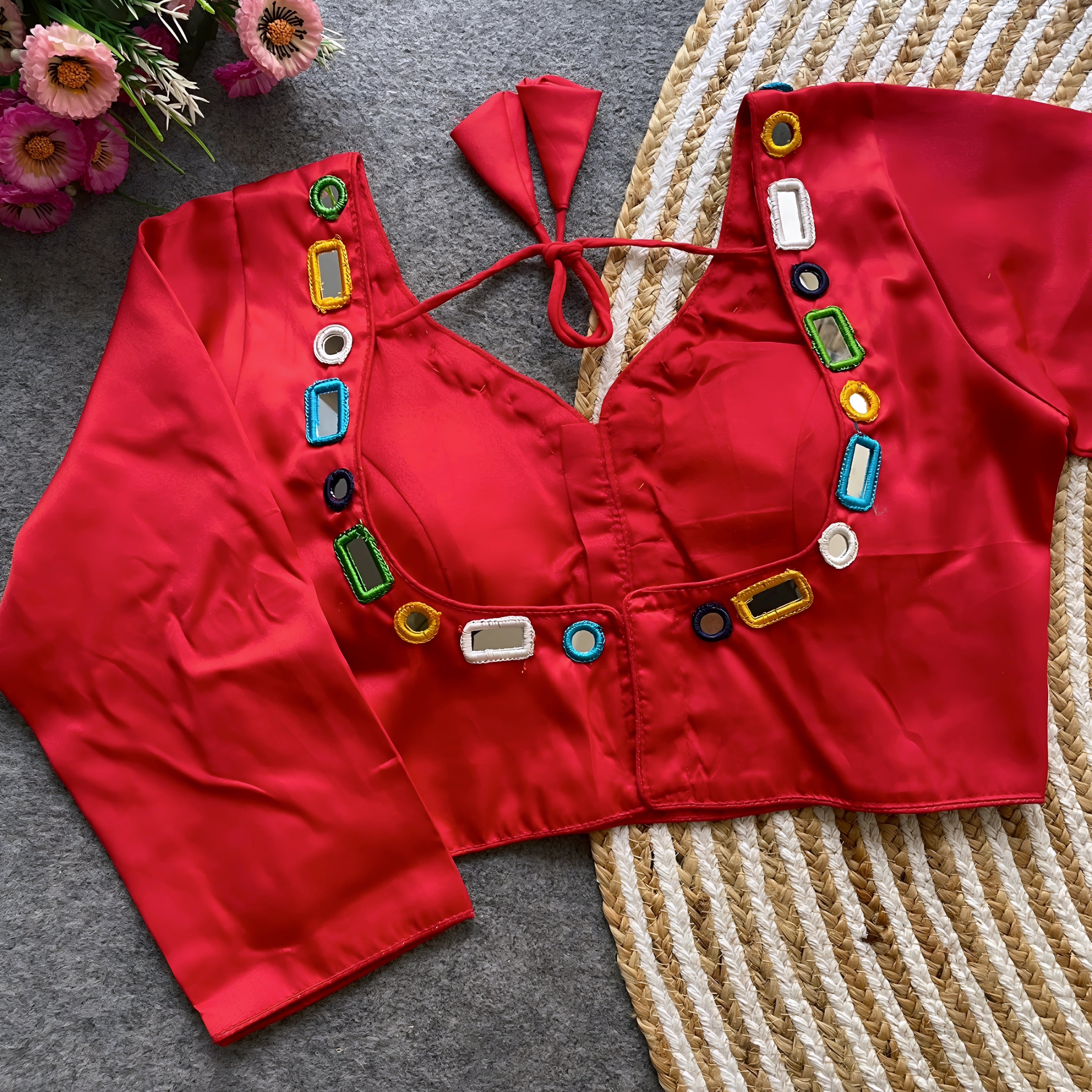 Red blouse embellished with square and round mirror work, featuring multicolored embroidered borders, stylish back tie-up closure, and three-quarter sleeves. Displayed flat on a textured grey surface alongside a woven mat and fresh flowers, highlighting its festive and contemporary ethnic style.