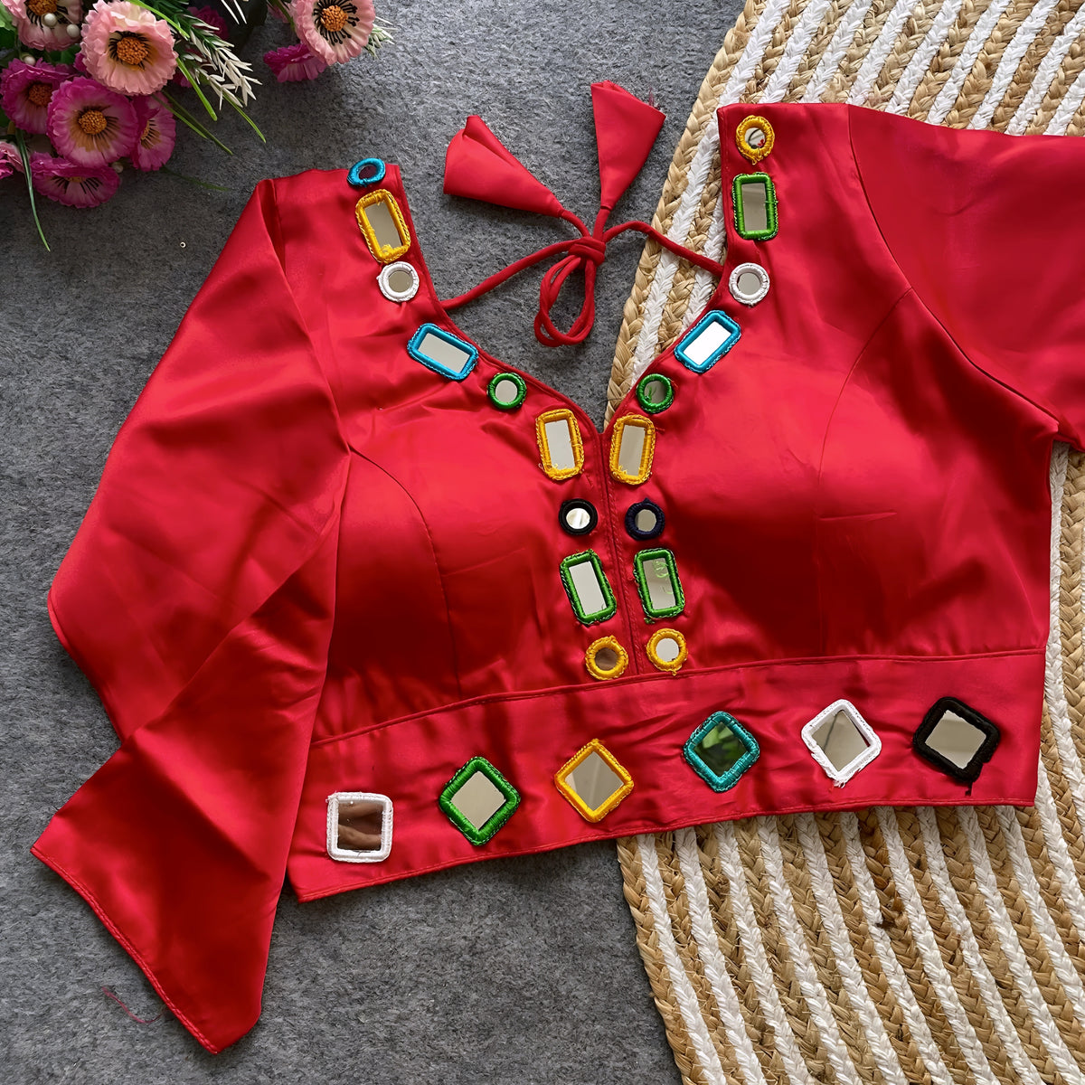Red blouse embellished with square and round mirror work, featuring multicolored embroidered borders, stylish back tie-up closure, and three-quarter sleeves. Displayed flat on a textured grey surface alongside a woven mat and fresh flowers, highlighting its festive and contemporary ethnic style.