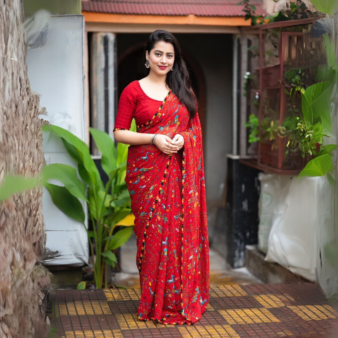 Woman in a red Warli print saree with pom-pom border standing on a tiled patio with lush greenery and a traditional house in the background
