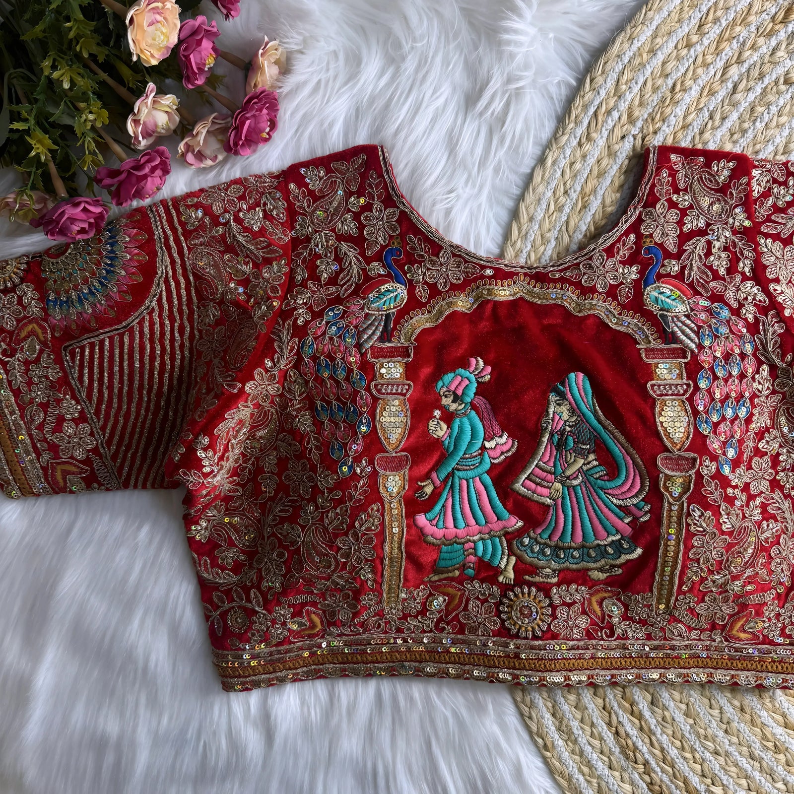 Traditional red silk blouse featuring intricate gold zardosi embroidery with floral and leaf motifs, sequin polka dots, striped geometric panels, and a multicolor peacock feather design on the sleeve, displayed flat on a white fur surface with a braided mat and artificial pink flowers in the corner.