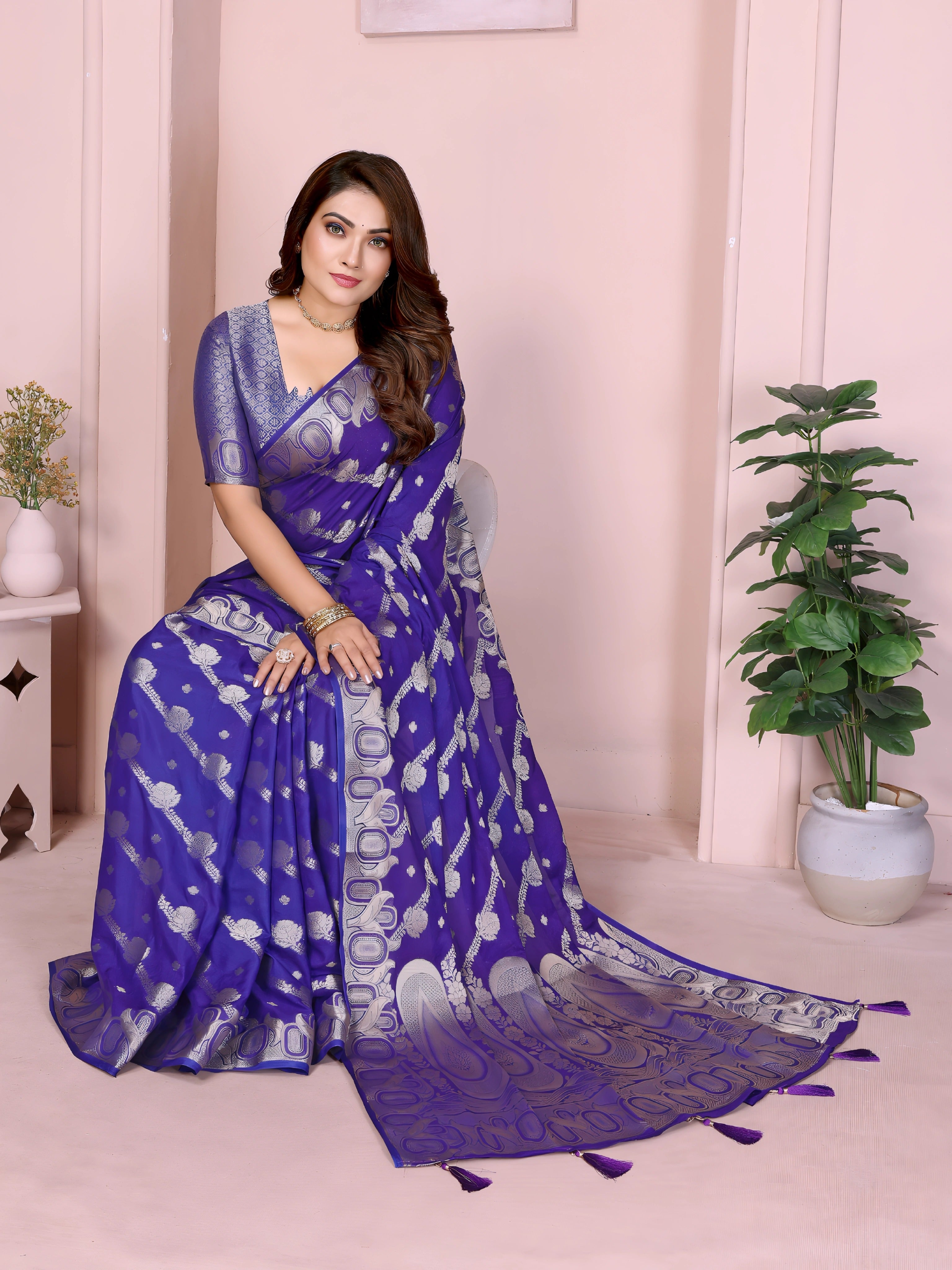 Woman wearing a royal blue Banarasi saree decorated with intricate silver zari motifs and an ornate border with tassel details, paired with a matching textured blue blouse and styled with traditional jewelry and bangles. She poses gracefully against a pastel arched backdrop, featuring minimalist decor including a white pedestal with vases and a potted green plant, highlighting an elegant festive ethnic look.
