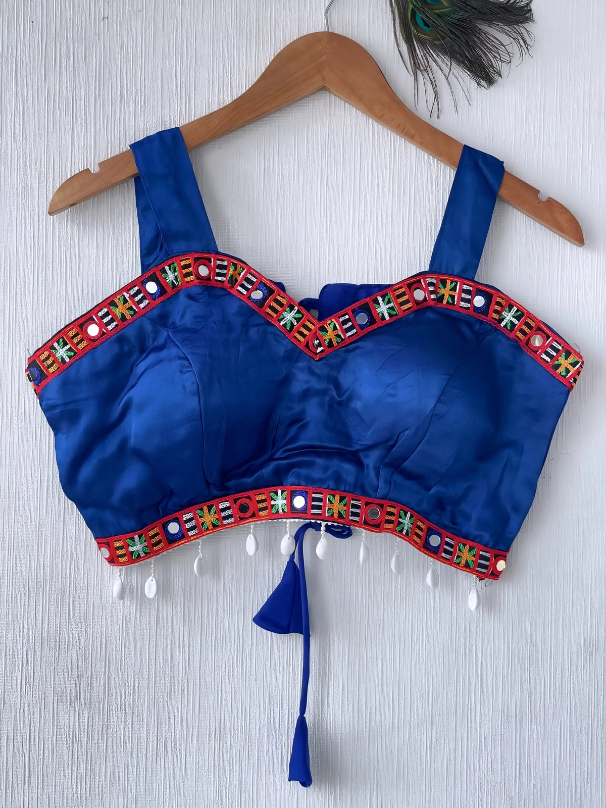  Royal blue ethnic blouse featuring vibrant mirror work and a multicolored embroidered geometric border in red, green, white, and orange, adorned with white pearl drop tassels and a tie-up waist detail. Displayed on a wooden hanger against a textured off-white wall with peacock feather, creating a festive and traditional look.