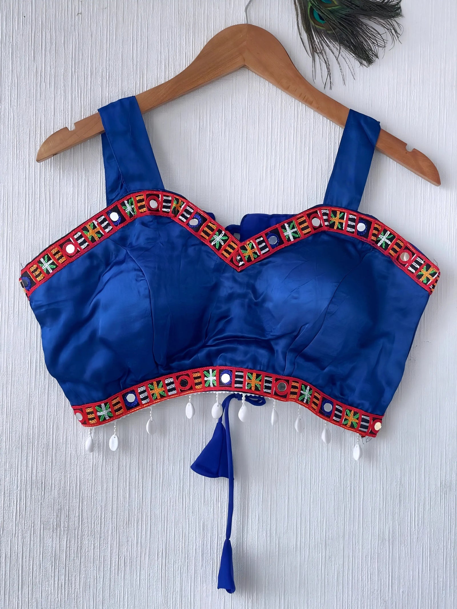  Royal blue ethnic blouse featuring vibrant mirror work and a multicolored embroidered geometric border in red, green, white, and orange, adorned with white pearl drop tassels and a tie-up waist detail. Displayed on a wooden hanger against a textured off-white wall with peacock feather, creating a festive and traditional look.