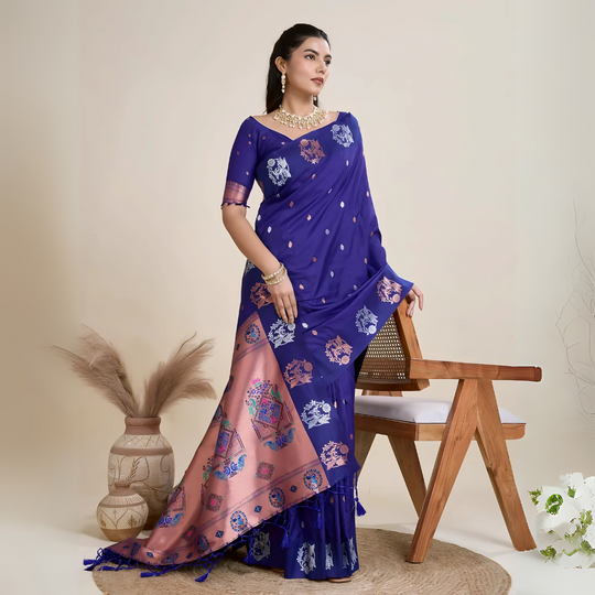 A woman models a luxurious Royal Blue Soft Silk Saree. The saree features silver Zari motifs on the body and a wide pallu in contrasting rose gold/peach, richly woven with multi-colored peacock and floral designs. She wears a matching short-sleeve blue blouse and heavy gold Kundan jewelry, posing indoors.