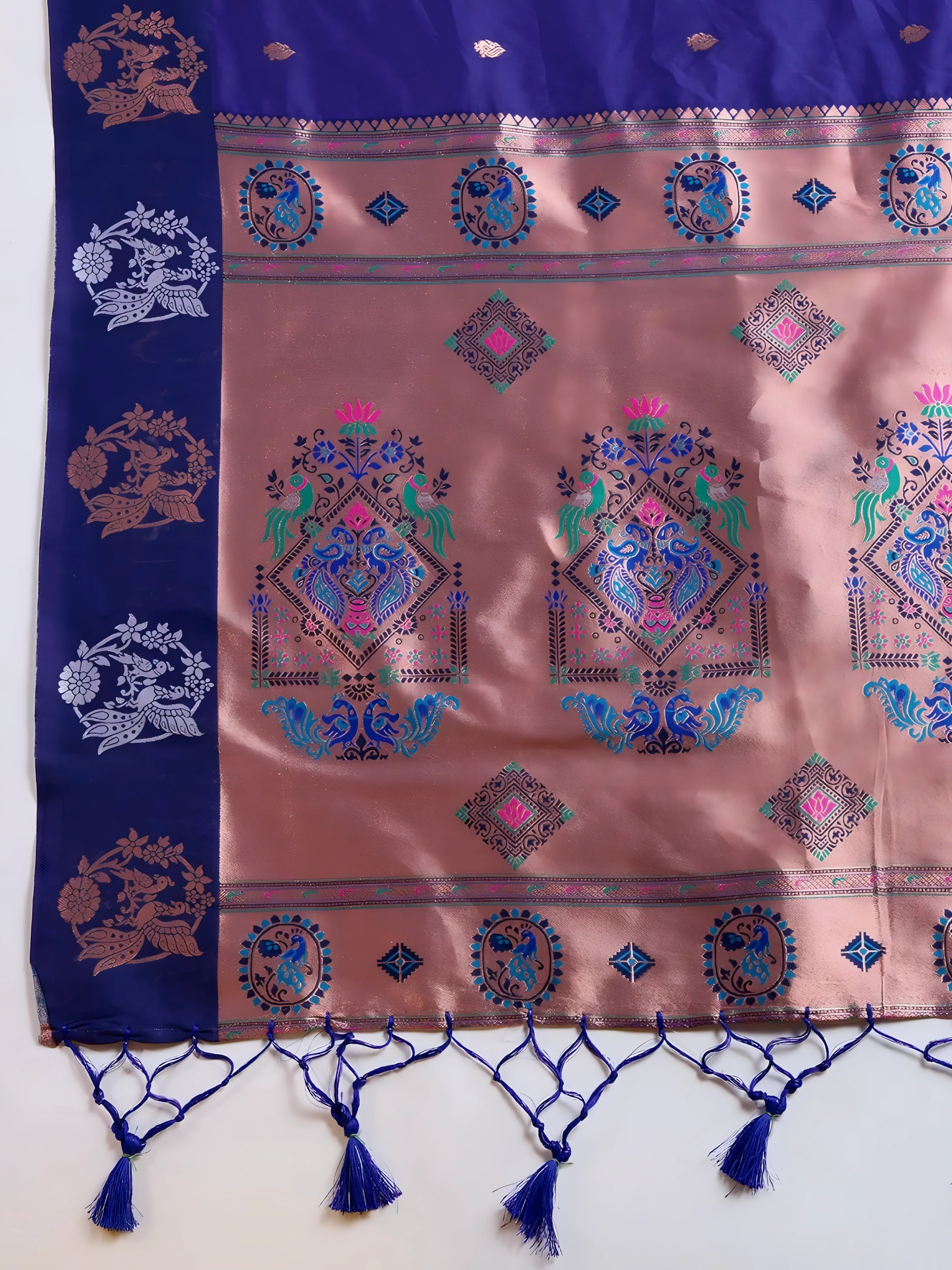 A woman models a luxurious Royal Blue Soft Silk Saree. The saree features silver Zari motifs on the body and a wide pallu in contrasting rose gold/peach, richly woven with multi-colored peacock and floral designs. She wears a matching short-sleeve blue blouse and heavy gold Kundan jewelry, posing indoors.