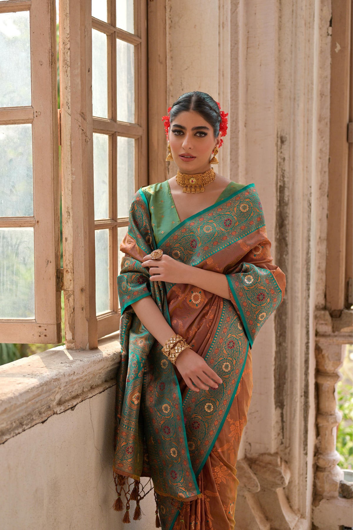 A woman models a striking Bronze Brown Banarasi Silk Saree with all-over Zari weaving. The saree has a wide, ornate Sea Green/Teal border featuring traditional Zari weaving in gold and rust tones. She wears a mint green blouse and gold Kundan jewelry, posing by a large antique window.