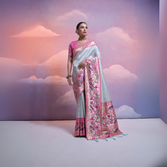 A woman models an elegant Light Blue/Icy Gray Soft Banarasi Silk Saree. The saree features a wide, vibrant Fuchsia Pink border and pallu with dense, colorful floral Kanjivaram-style weaving. She wears a contrasting short-sleeve pink blouse and a gold choker necklace, posing indoors against a surreal pink and blue cloud backdrop.