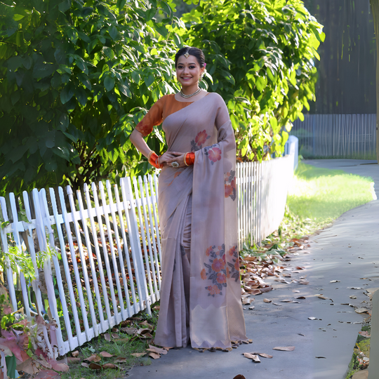 A woman models an elegant Dusty Gray Soft Muga Cotton Saree. The pallu features large, artistic Resham floral weaving in pink and coral tones. She wears a contrasting rust orange blouse and is accessorized with a pearl choker necklace and vibrant orange bangles, posing outdoors by a white fence.