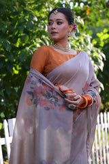 A woman models an elegant Dusty Gray Soft Muga Cotton Saree. The pallu features large, artistic Resham floral weaving in pink and coral tones. She wears a contrasting rust orange blouse and is accessorized with a pearl choker necklace and vibrant orange bangles, posing outdoors by a white fence.
