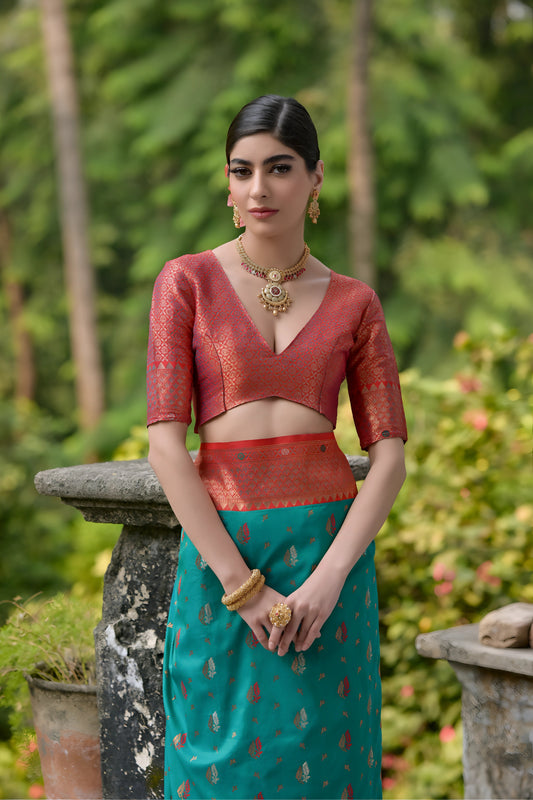 A woman models a luxurious Teal Banarasi Silk Saree with scattered gold Zari motifs. The wide border and pallu contrast dramatically in Terracotta Red with dense traditional gold weaving. She wears a coral short-sleeve blouse and heavy gold Kundan jewelry, posing on an ornate stone terrace.