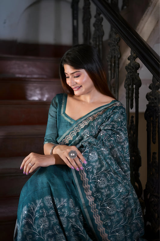 A woman models an elegant Deep Teal Soft Tussar Silk Saree. The saree features dense all-over floral embroidery/weaving in a light metallic thread, framed by a contrasting light metallic border with geometric patterns. She wears a matching teal blouse and large oxidized silver earrings, posing indoors by a vibrant green door.