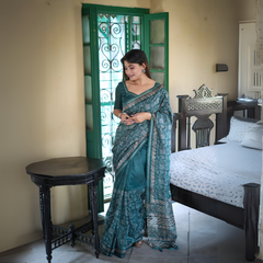 A woman models an elegant Deep Teal Soft Tussar Silk Saree. The saree features dense all-over floral embroidery/weaving in a light metallic thread, framed by a contrasting light metallic border with geometric patterns. She wears a matching teal blouse and large oxidized silver earrings, posing indoors by a vibrant green door.