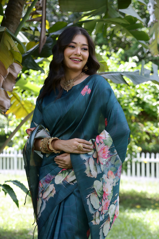 A woman models a stunning Deep Rama Blue Soft Tussar Silk Saree. The saree features a large-scale digital print of colorful floral and lotus motifs in pink, white, and green on the pallu and lower drape. She wears a matching elbow-sleeve blue blouse and heavy gold Kundan jewelry, posing outdoors in a garden setting.