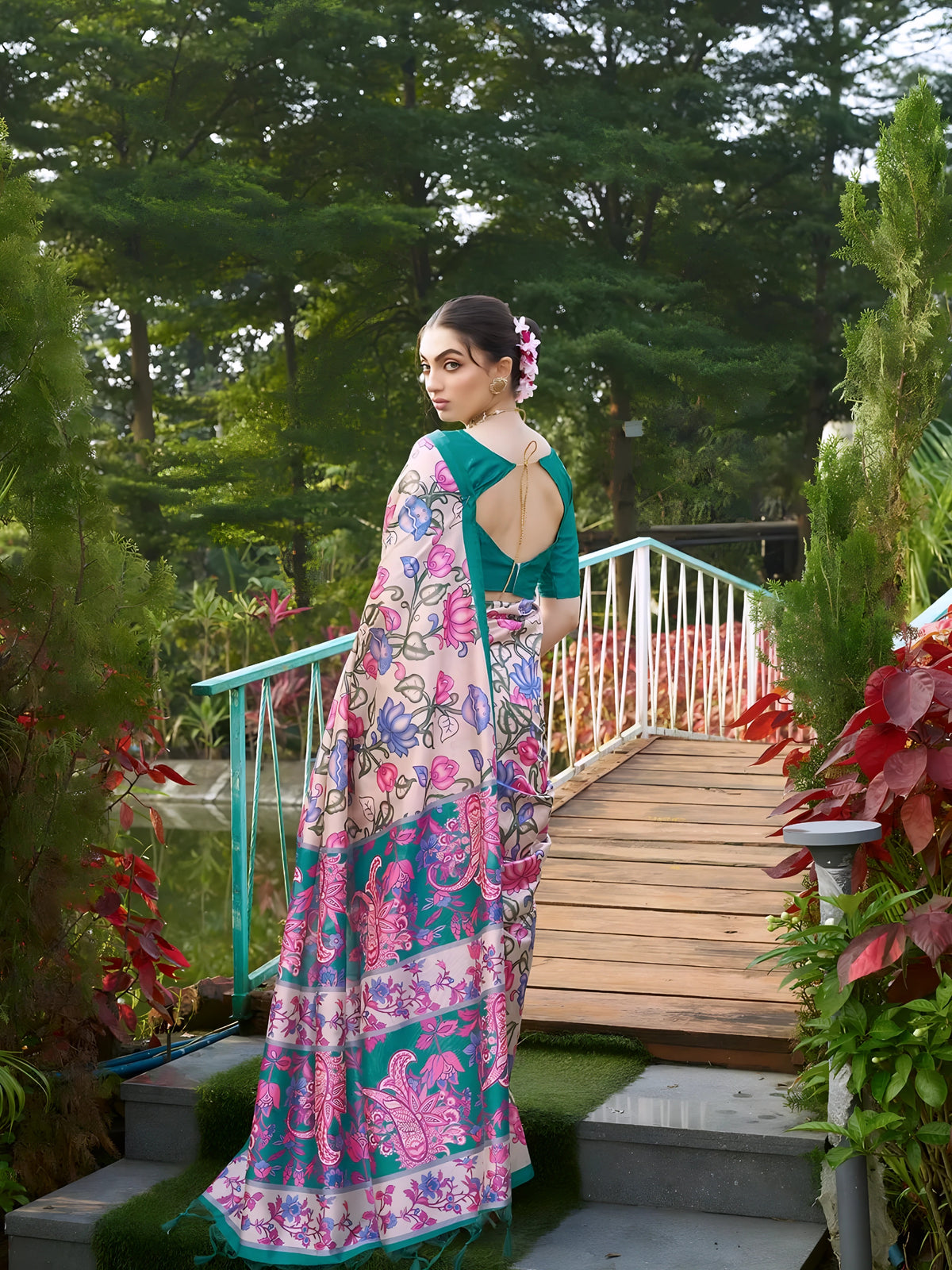 A woman models an elegant Beige Soft Tussar Silk Saree with a dense all-over print of pink, blue, and purple floral and vine motifs. The saree has a wide, contrasting Deep Teal border and pallu with traditional weaving/print. She wears a matching teal blouse and a gold Kundan choker necklace, posing outdoors on a wooden bridge.
