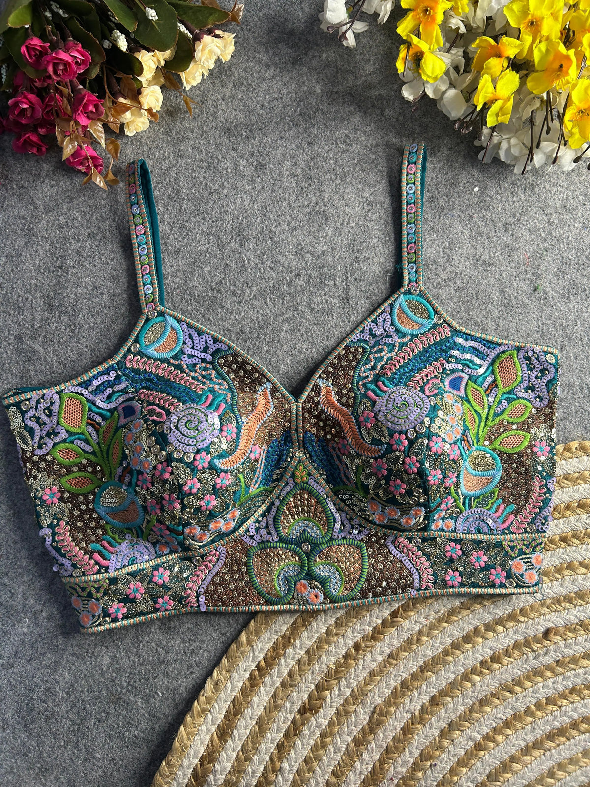Traditional teal bustier-style blouse featuring vibrant multicolor Madhubani-inspired embroidery with intricate floral, paisley, and geometric motifs, embellished with sequins and beads, structured cups, and adjustable straps, displayed flat on a textured grey surface with artificial pink, yellow, and white flowers in the corners.
