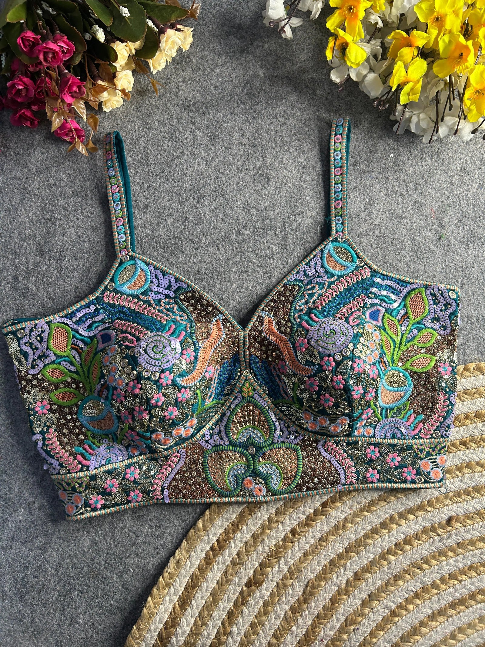 Traditional teal bustier-style blouse featuring vibrant multicolor Madhubani-inspired embroidery with intricate floral, paisley, and geometric motifs, embellished with sequins and beads, structured cups, and adjustable straps, displayed flat on a textured grey surface with artificial pink, yellow, and white flowers in the corners.