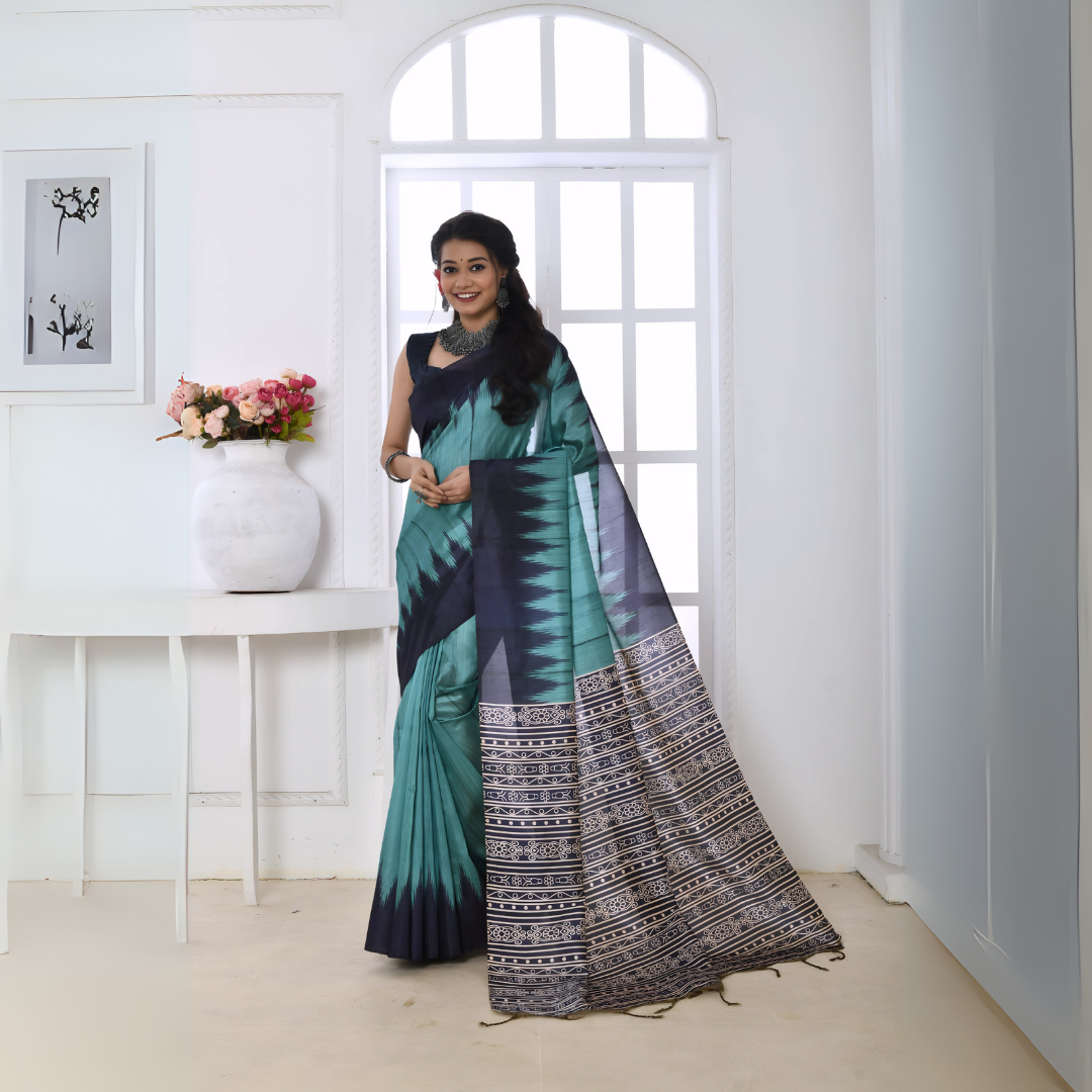 A woman models a vibrant Sky Blue/Teal Soft Tussar Silk Saree. The saree features a bold black/navy zigzag pattern along the border and transitions on the body. The pallu has a wide panel of dense tribal or Ikat-style geometric print in light grey, black, and white. She wears a dark sleeveless blouse and chunky oxidized silver jewelry, posing indoors by a large arched window.