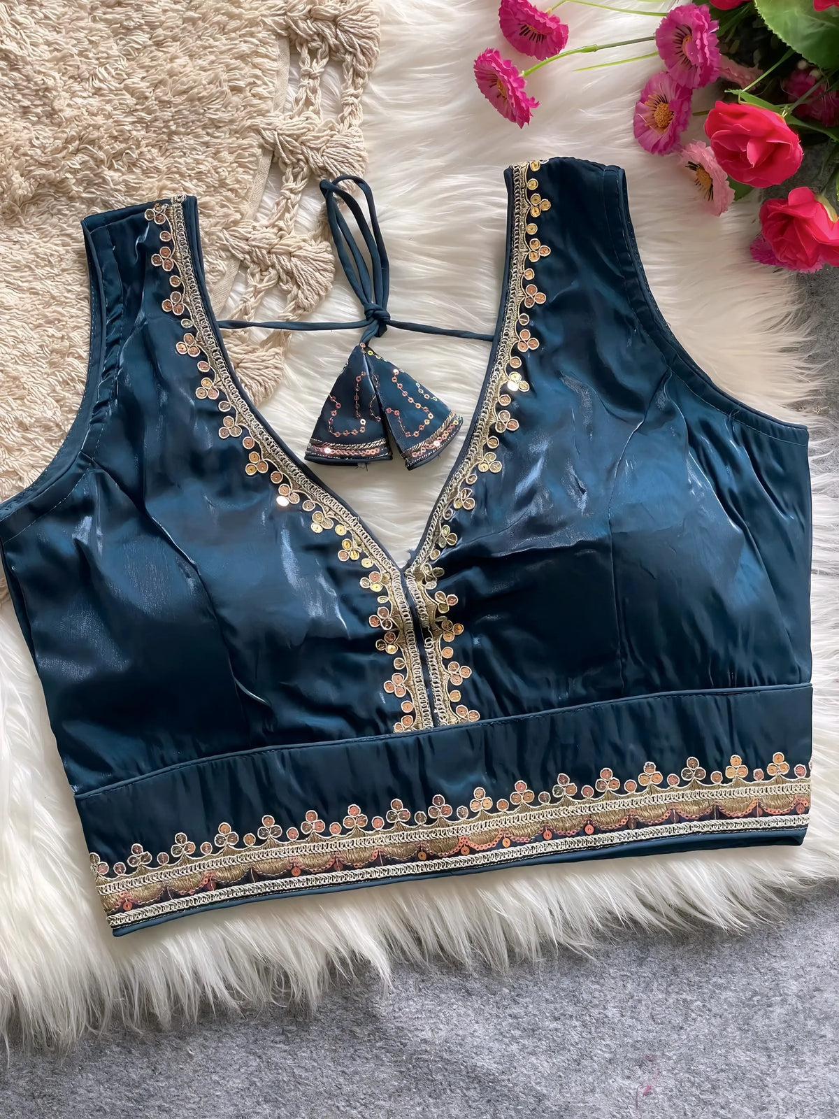 Traditional teal silk blouse featuring intricate gold sequin and thread embroidery along the neckline and hem, with decorative tassel tie at the back, displayed flat on a soft fur and textured mat background with artificial pink flowers in the corner.