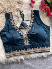 Traditional teal silk blouse featuring intricate gold sequin and thread embroidery along the neckline and hem, with decorative tassel tie at the back, displayed flat on a soft fur and textured mat background with artificial pink flowers in the corner.