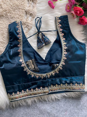 Traditional teal silk blouse featuring intricate gold sequin and thread embroidery along the neckline and hem, with decorative tassel tie at the back, displayed flat on a soft fur and textured mat background with artificial pink flowers in the corner.