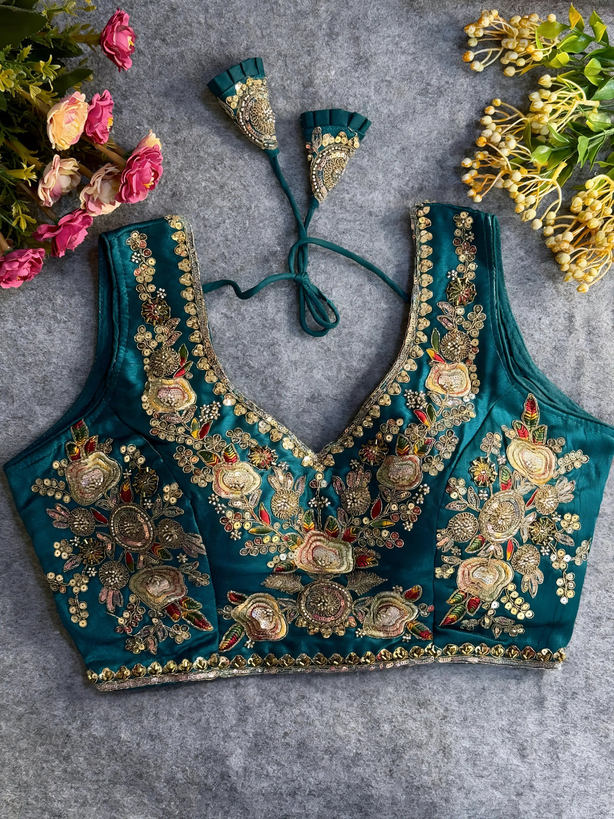 Traditional teal silk blouse featuring intricate gold zardozi embroidery and beadwork with floral and leaf motifs on the front and neckline, finished with decorative tassel ties, displayed flat on a textured grey background with artificial pink and yellow flowers in the corners.