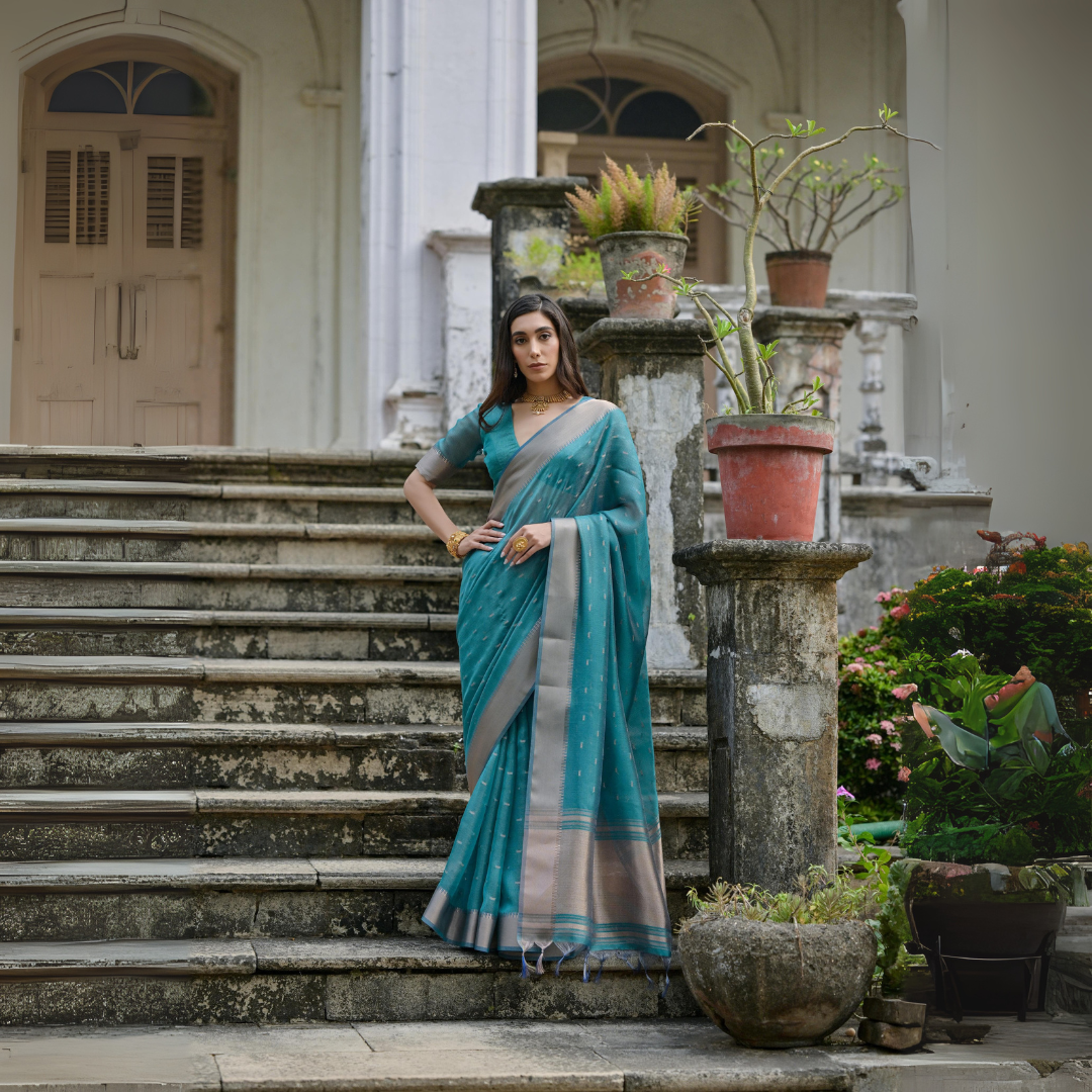 A woman models a stunning Deep Sea Blue Soft Silk Saree. The saree features scattered metallic motifs and a wide, contrasting silver/gray Zari woven border. She wears a matching blue elbow-sleeve blouse and a delicate gold choker necklace, posing on an ornate stone staircase.