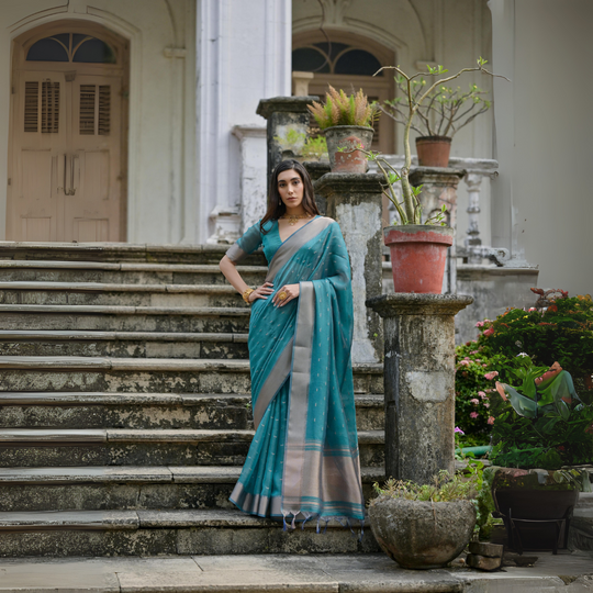 A woman models a stunning Deep Sea Blue Soft Silk Saree. The saree features scattered metallic motifs and a wide, contrasting silver/gray Zari woven border. She wears a matching blue elbow-sleeve blouse and a delicate gold choker necklace, posing on an ornate stone staircase.