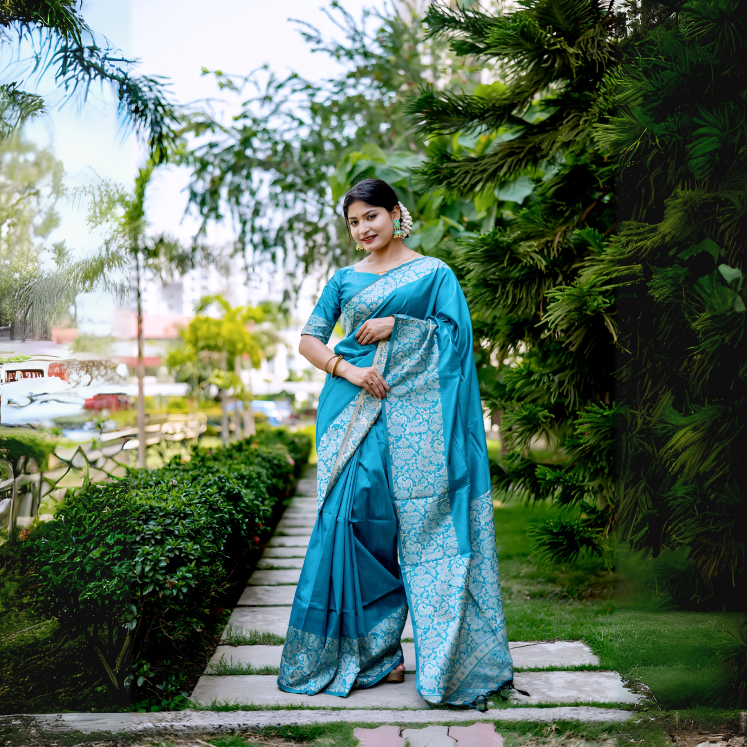A woman models a luxurious Aqua Blue Raw Silk Saree. The saree features a wide border and pallu with dense, intricate silver Zari weaving in a Kalamkari-style floral and paisley pattern. She wears a matching short-sleeve blue blouse and subtle metallic jewelry, with white flowers adorning her hair, posing outdoors on a stone pathway.