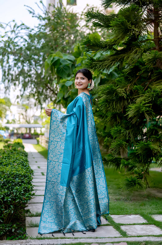 A woman models a luxurious Aqua Blue Raw Silk Saree. The saree features a wide border and pallu with dense, intricate silver Zari weaving in a Kalamkari-style floral and paisley pattern. She wears a matching short-sleeve blue blouse and subtle metallic jewelry, with white flowers adorning her hair, posing outdoors on a stone pathway.
