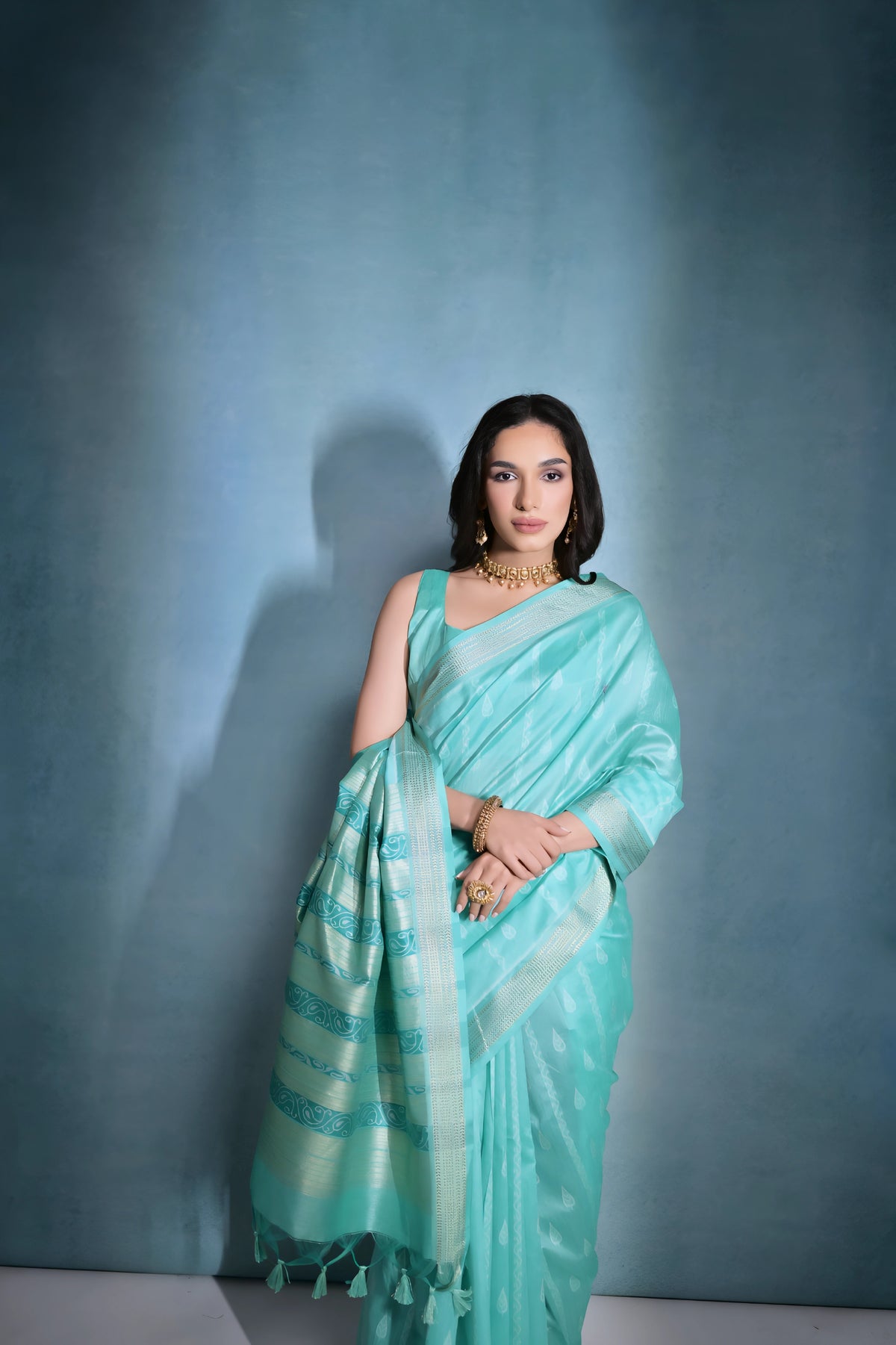 A woman models a serene Sky Blue Soft Raw Silk Saree. The saree body features subtle tone-on-tone Ikat weaving. The pallu is highlighted by wide horizontal bands of metallic silver/gold Zari weaving. She wears a blue sleeveless blouse and gold Kundan jewelry.