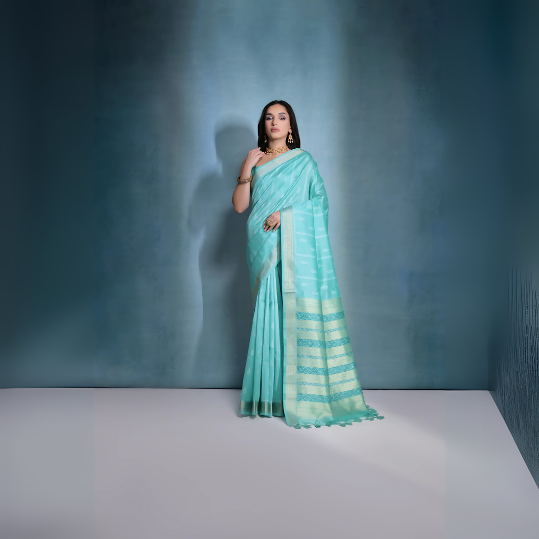 A woman models a serene Sky Blue Soft Raw Silk Saree. The saree body features subtle tone-on-tone Ikat weaving. The pallu is highlighted by wide horizontal bands of metallic silver/gold Zari weaving. She wears a blue sleeveless blouse and gold Kundan jewelry.