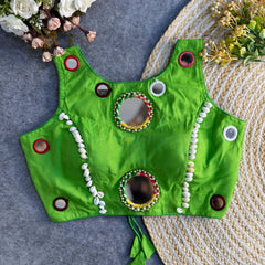 Bright green ethnic blouse with circular mirror work, cowrie shell borders, and colorful embroidery laying flat on a woven mat