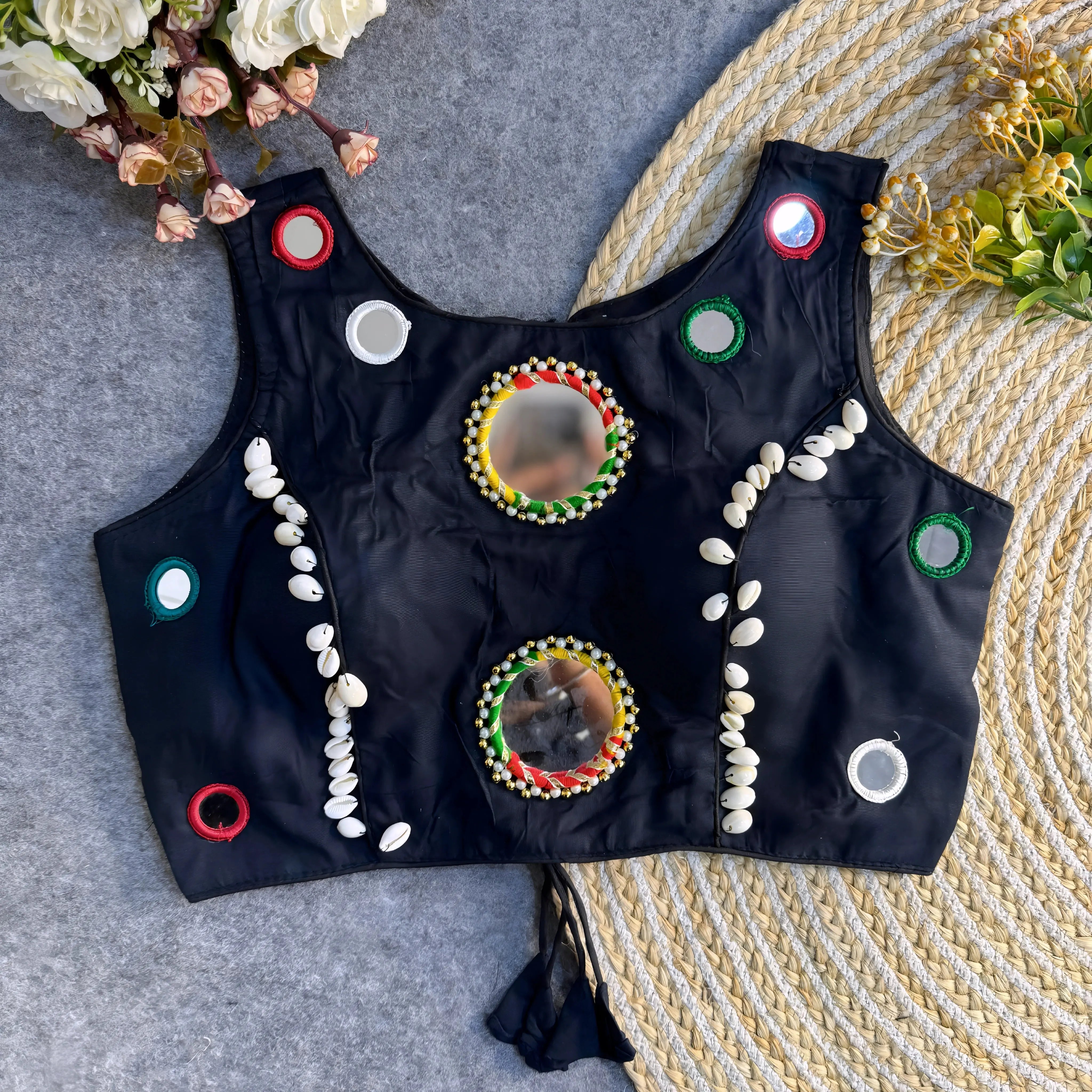Jet-black Indian blouse embellished with multicolor mirror circles and rows of cowrie shells, positioned flat on grey fabric beside a braided jute rug.