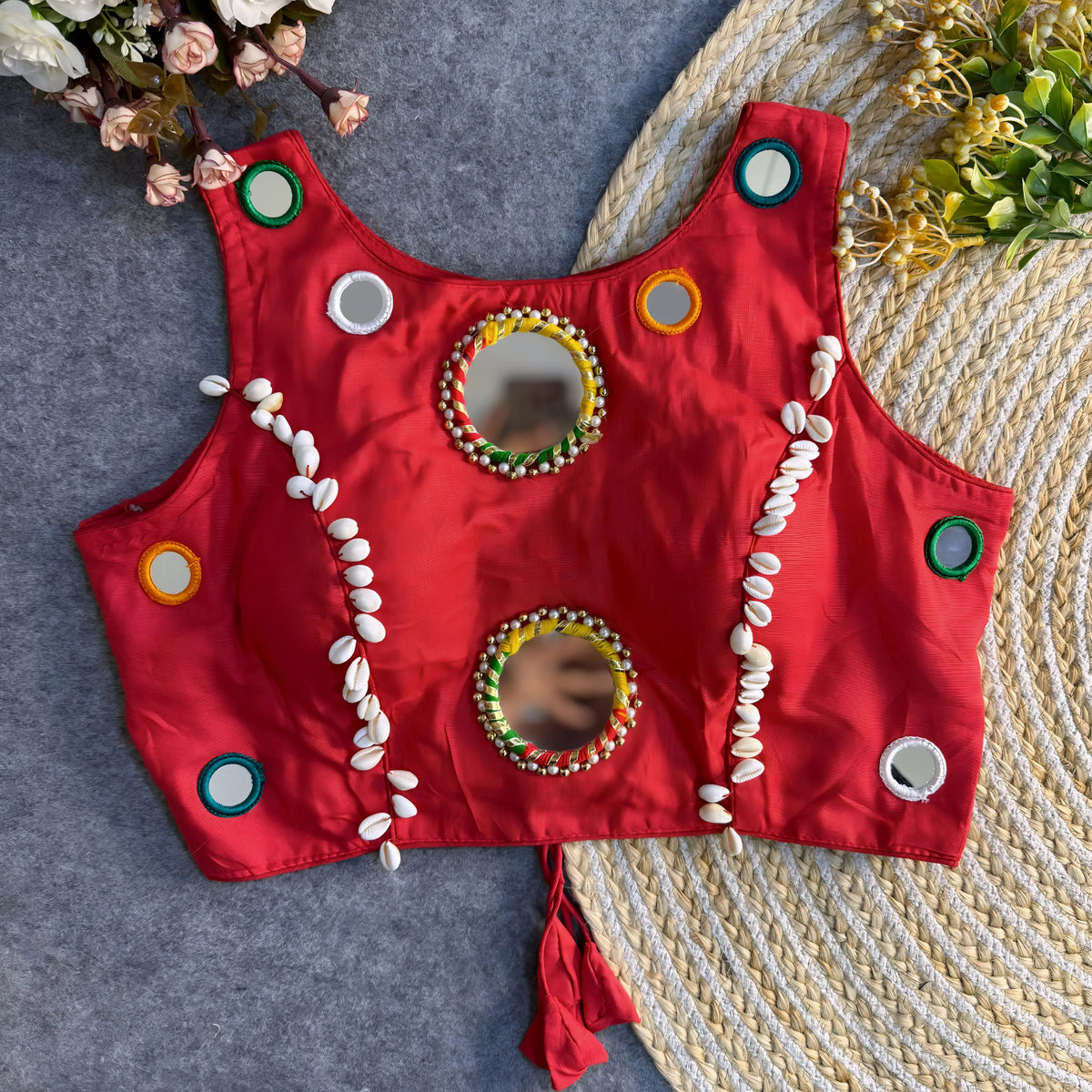 Crimson red Indian blouse featuring mirror embroidery and cowrie shell accents, arranged flat on grey fabric and jute rug.