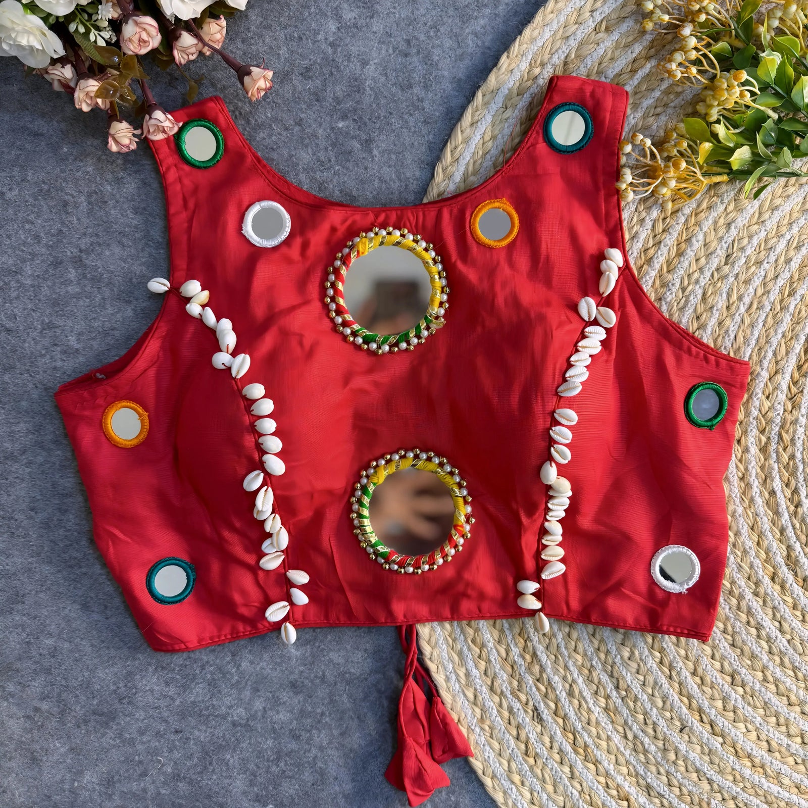 Crimson red Indian blouse featuring mirror embroidery and cowrie shell accents, arranged flat on grey fabric and jute rug.