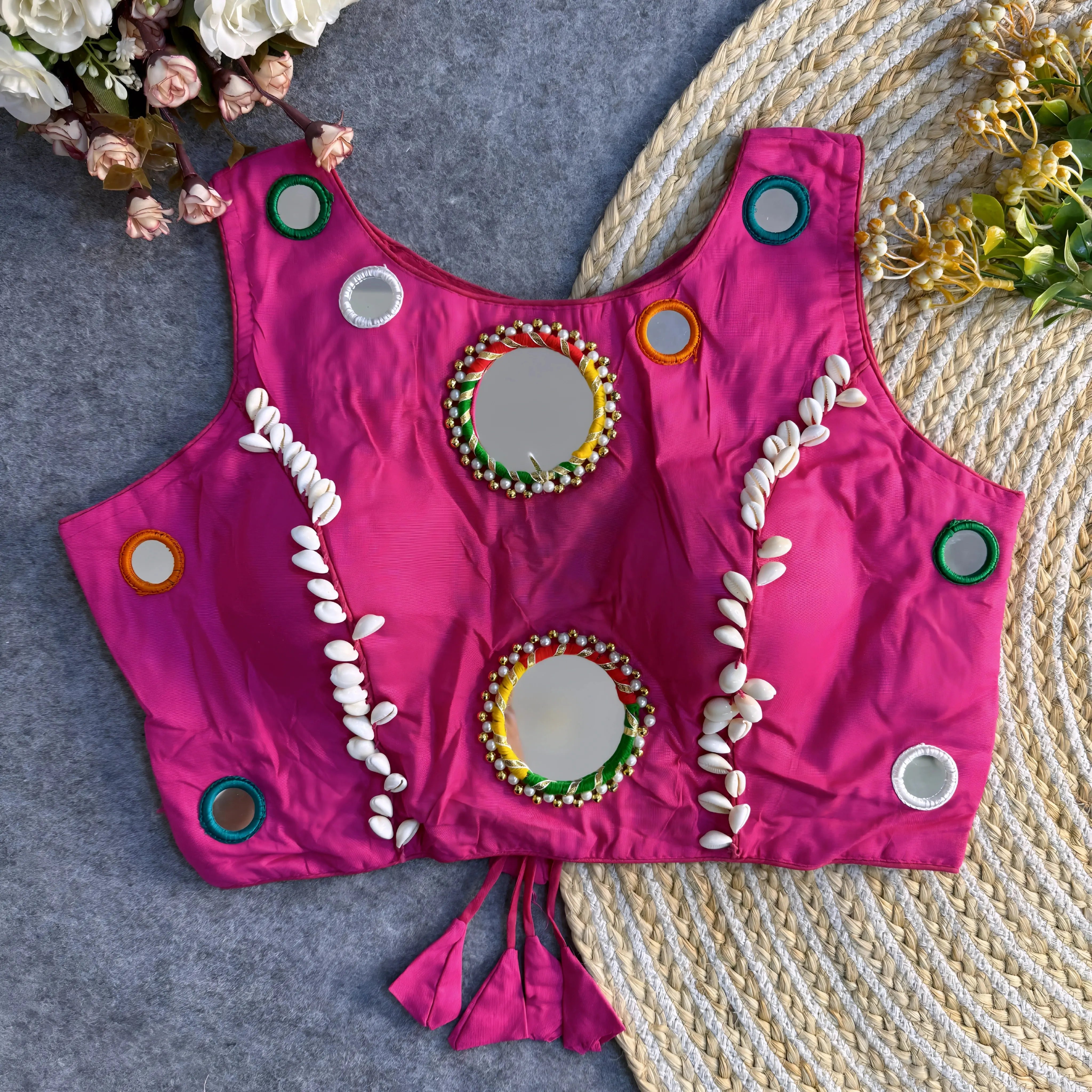 Bright fuchsia Indian blouse adorned with round mirrors and cowrie shell embellishments laid flat on a textured background