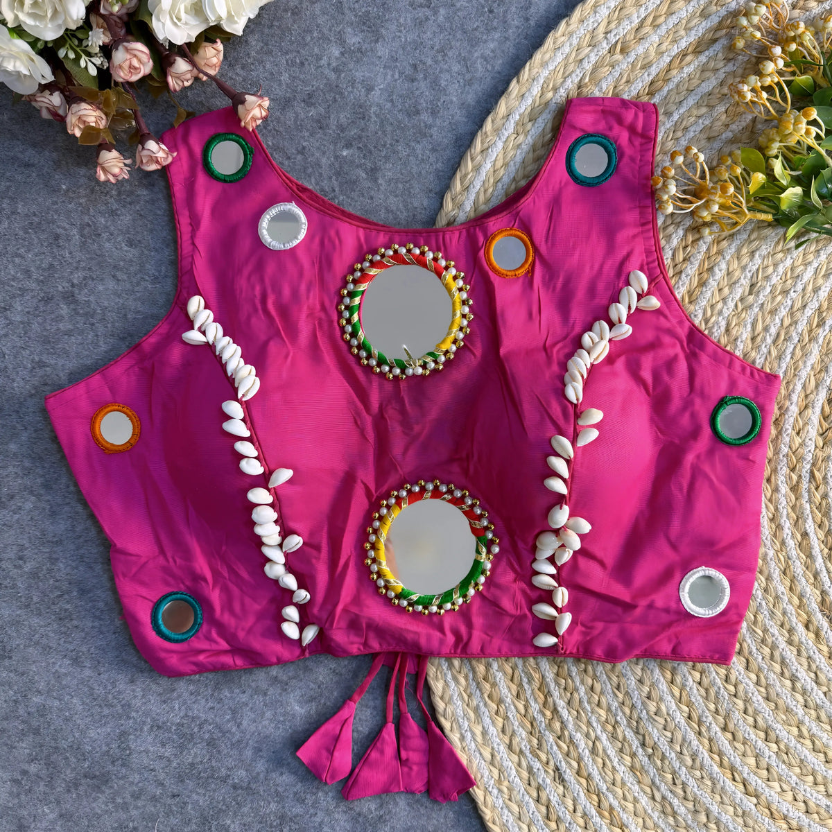 Bright fuchsia Indian blouse adorned with round mirrors and cowrie shell embellishments laid flat on a textured background