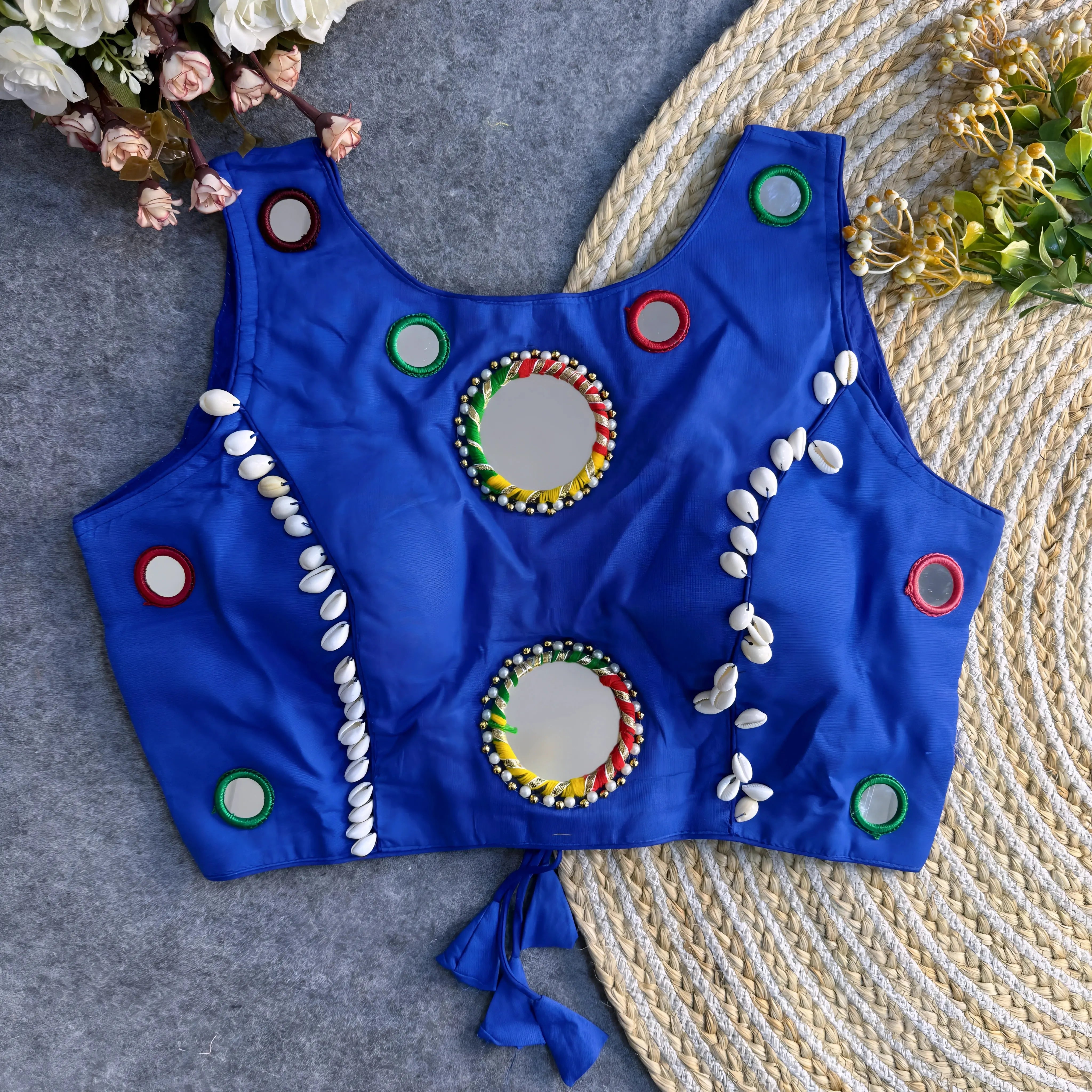 Royal-blue Indian blouse decorated with circular mirrors and cowrie shell designs, displayed flat on grey fabric and jute rug.