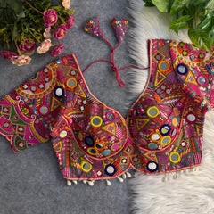 Traditional vibrant blouse featuring intricate multicolor mirror work and geometric embroidery in pink, yellow, green, orange, and blue, adorned with large and small round mirrors, beaded shell trim, and tasseled tie-back, displayed flat on a grey surface with artificial pink flowers and lush green foliage at the corners.