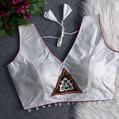  White padded blouse with red piping, featuring a central embroidered triangle motif with mirror work and shell detailing along the hem, displayed on a grey surface with artificial pink flowers and white fur in the background.